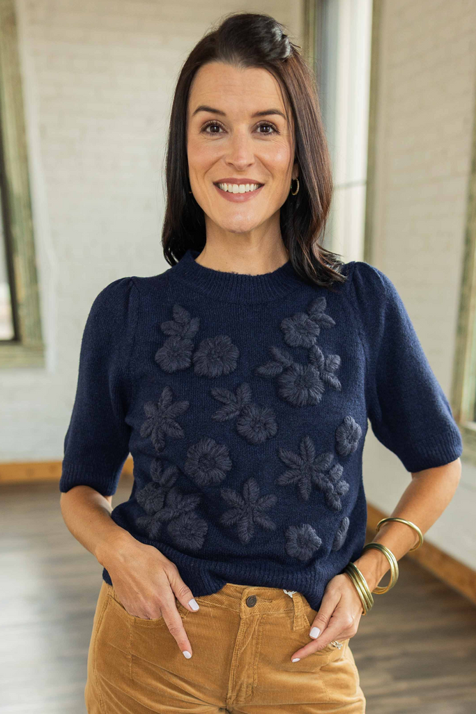 Kandace wearing a navy short sleeve knit top with floral appliqué detail on the front, paired with camel corduroy pants and brown sandals.