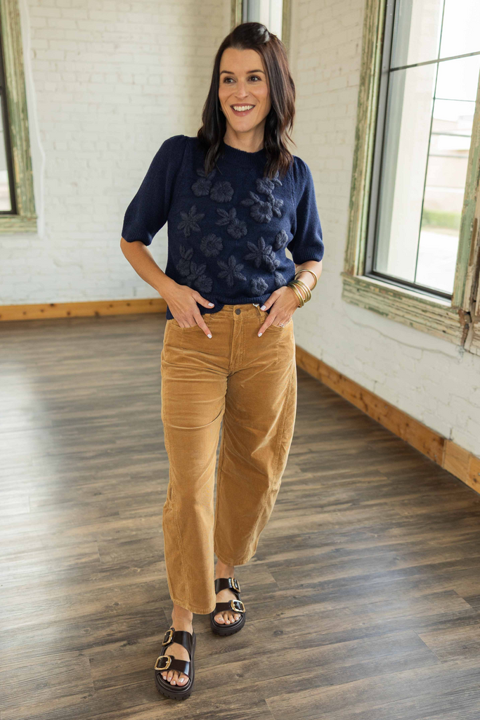 Kandace wearing a navy short sleeve knit top with floral appliqué detail on the front, paired with camel corduroy pants and brown sandals.