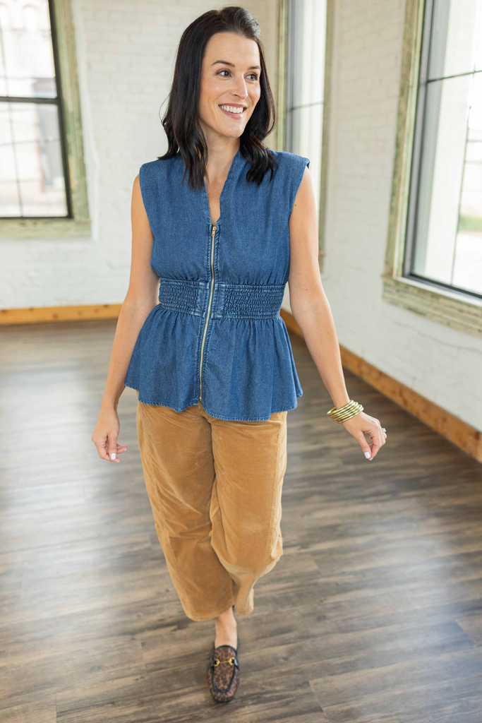 wearing a sleeveless denim peplum top with front zipper and smocked waist, paired with camel corduroy pants and loafers.