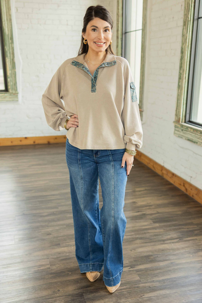 Kayla wearing a beige ribbed pullover with teal floral contrast trim, collared neckline, and button pocket on sleeve, paired with denim jeans.