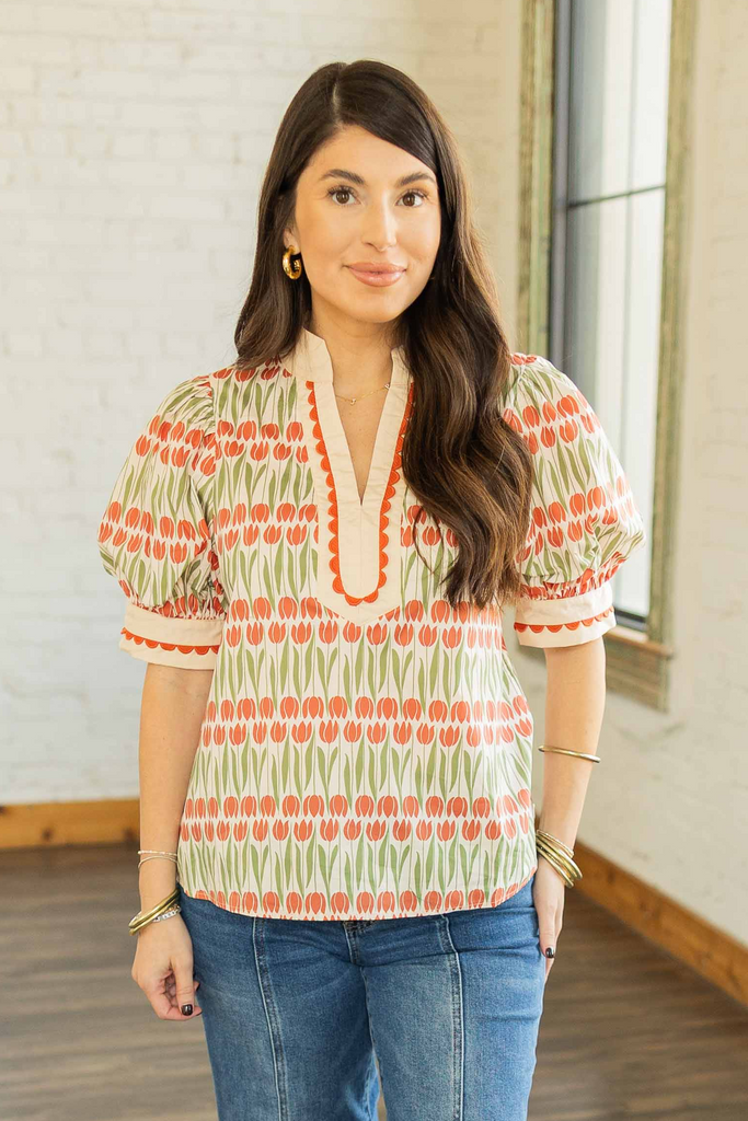 wearing a cream top with pumpkin orange and olive floral pattern, scalloped neckline trim, and short puff sleeves, paired with medium wash denim jeans.