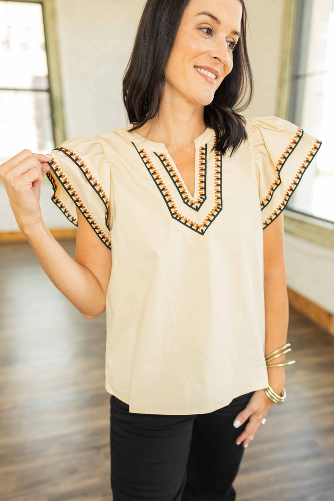 wearing a cream flutter sleeve top with black and copper embroidered trim around the V-neckline and sleeves, paired with black pants.