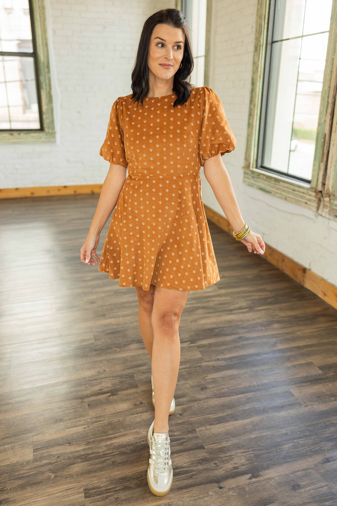 Kandace wearing amber-brown corduroy mini dress with cream floral print, short puff sleeves, and self-tie back detail paired with sneakers.