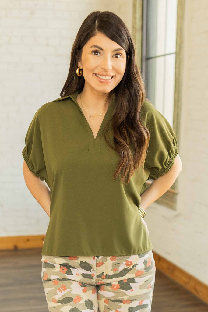 Kayla wearing an olive green short sleeve top with split collar and side slits, paired with floral camo wide-leg pants.
