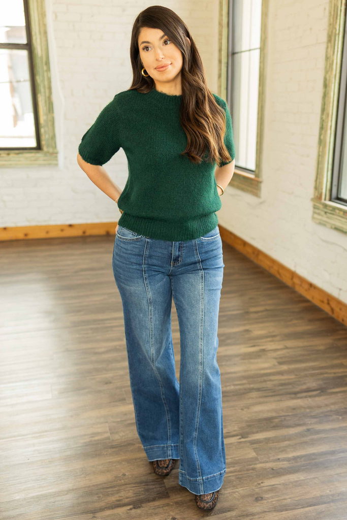 Kayla wearing a forest green short sleeve sweater knit top with a crew neckline, paired with medium wash denim jeans.