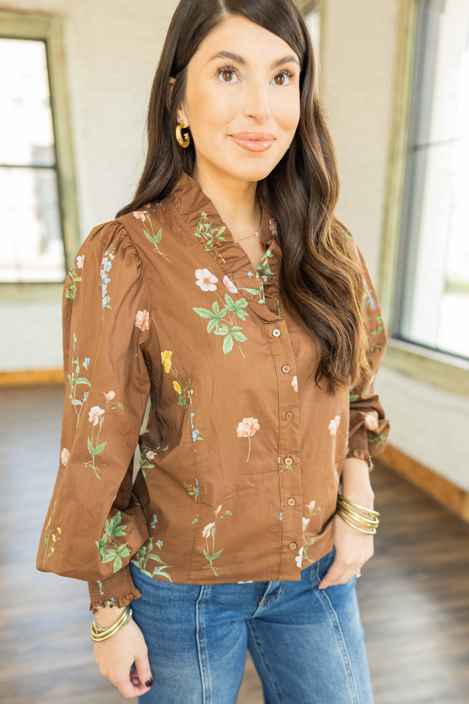 Kayla wearing a chestnut brown long sleeve floral top with smocked cuffs and ruffled neckline, paired with denim jeans.