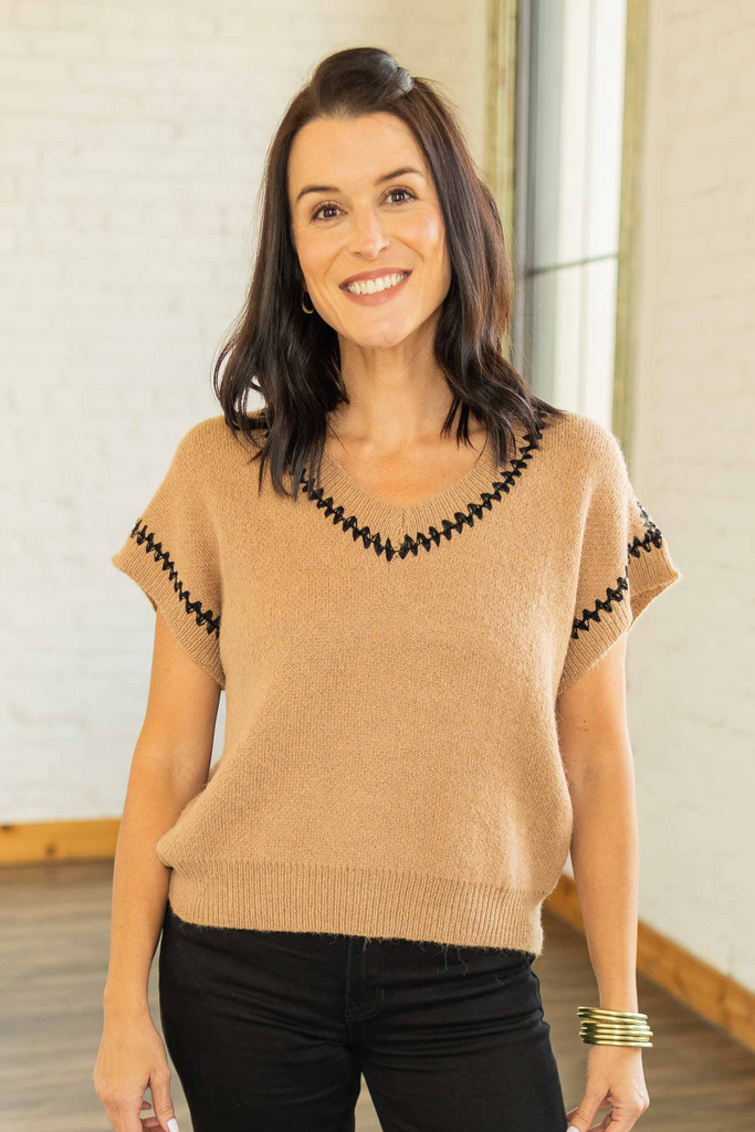 wearing a camel knit cap sleeve sweater top with black trim detail and V-neckline, paired with black cuffed pants and loafers.