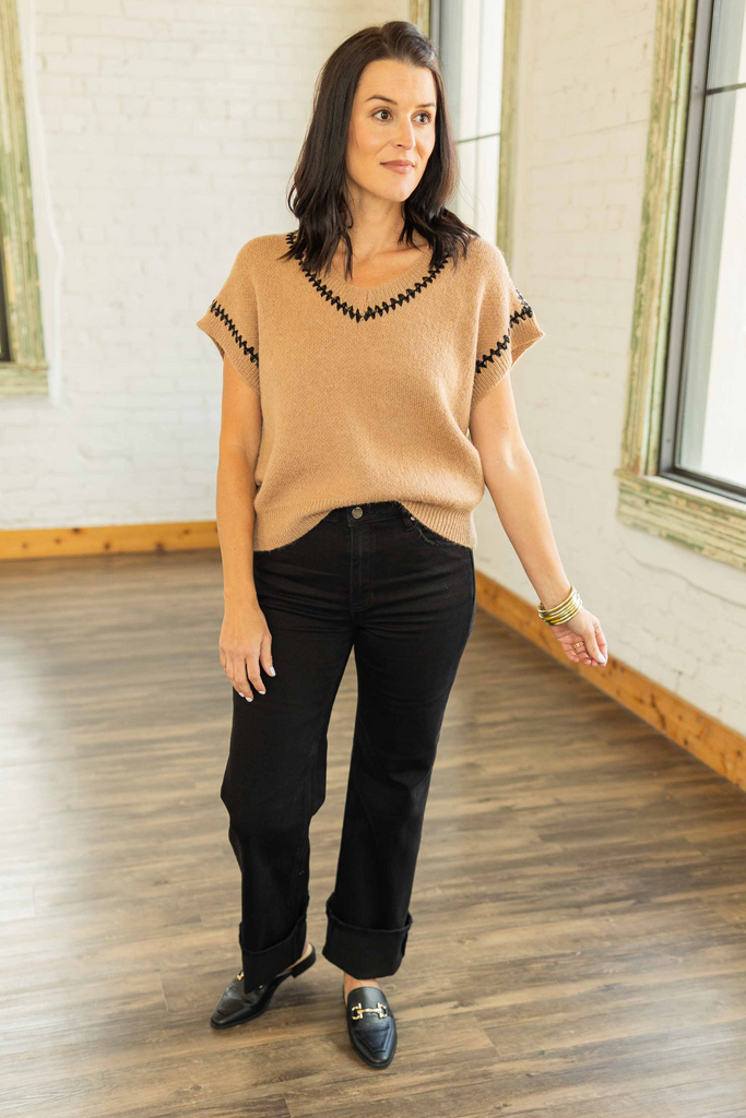 wearing a camel knit cap sleeve sweater top with black trim detail and V-neckline, paired with black cuffed pants and loafers.