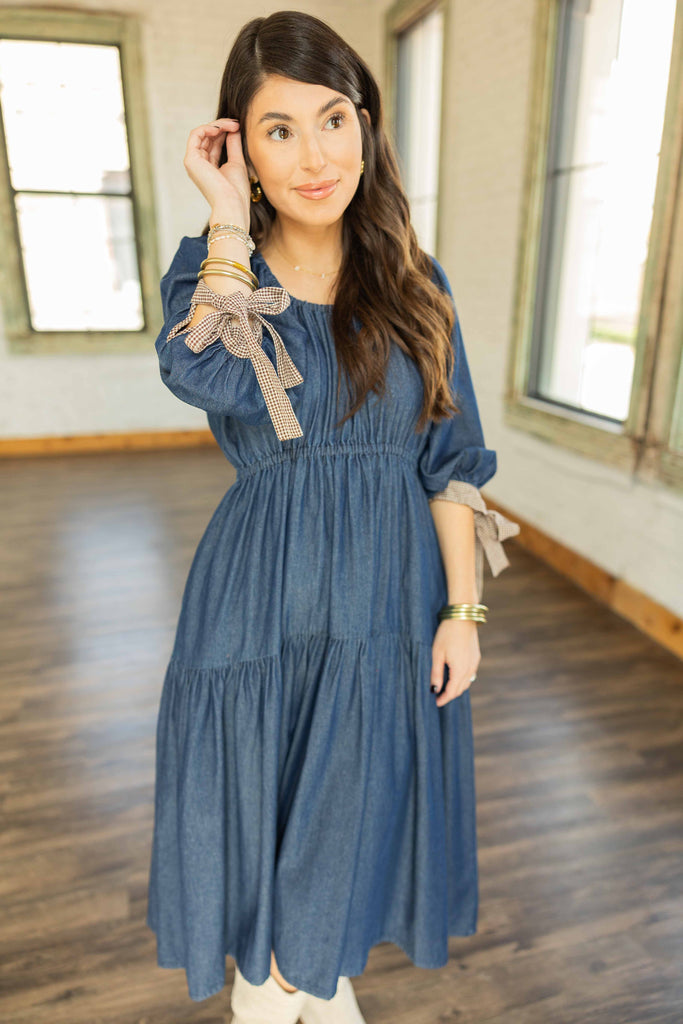 Kayla wearing tiered denim midi dress with elastic waist, 3/4 sleeves, and gingham tie cuffs paired with cream boots.