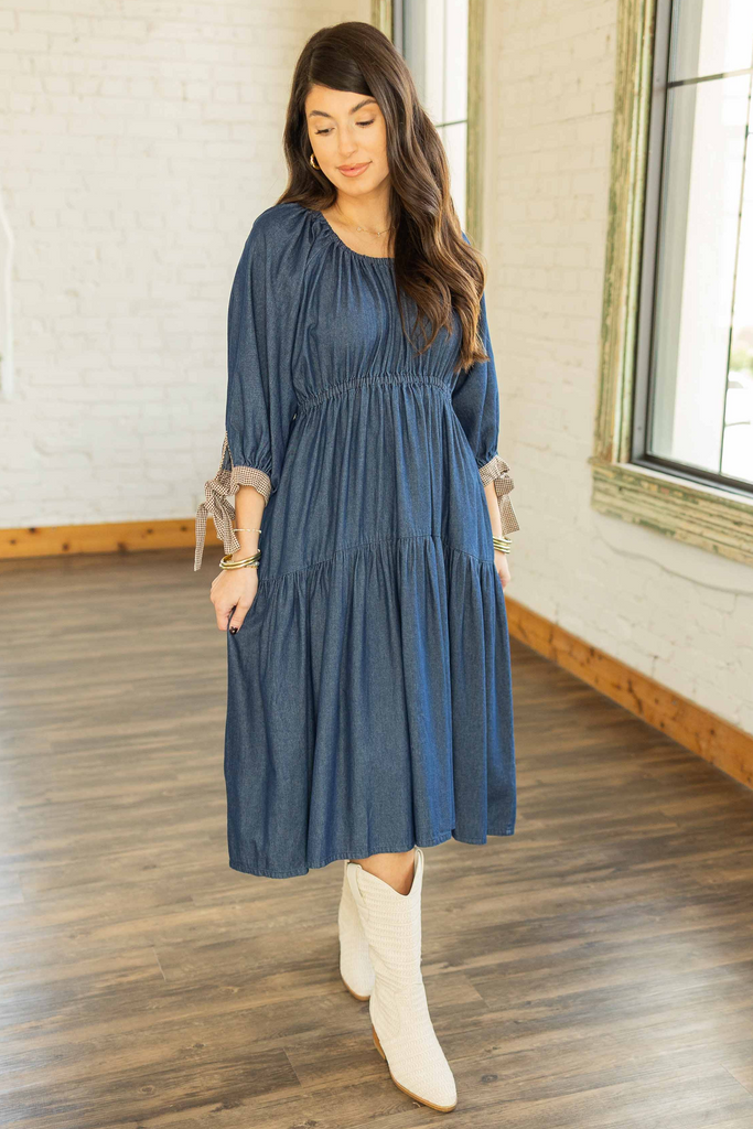 Kayla wearing tiered denim midi dress with elastic waist, 3/4 sleeves, and gingham tie cuffs paired with cream boots.