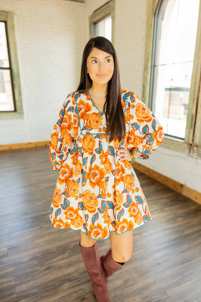 Kayla wearing the Noa cream mini dress with orange and teal floral print, rick-rack trim, and tall maroon boots