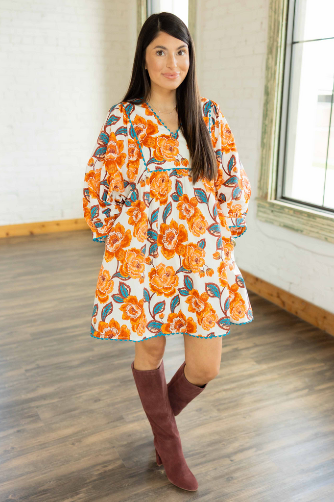 Kayla wearing the Noa cream mini dress with orange and teal floral print, rick-rack trim, and tall maroon boots