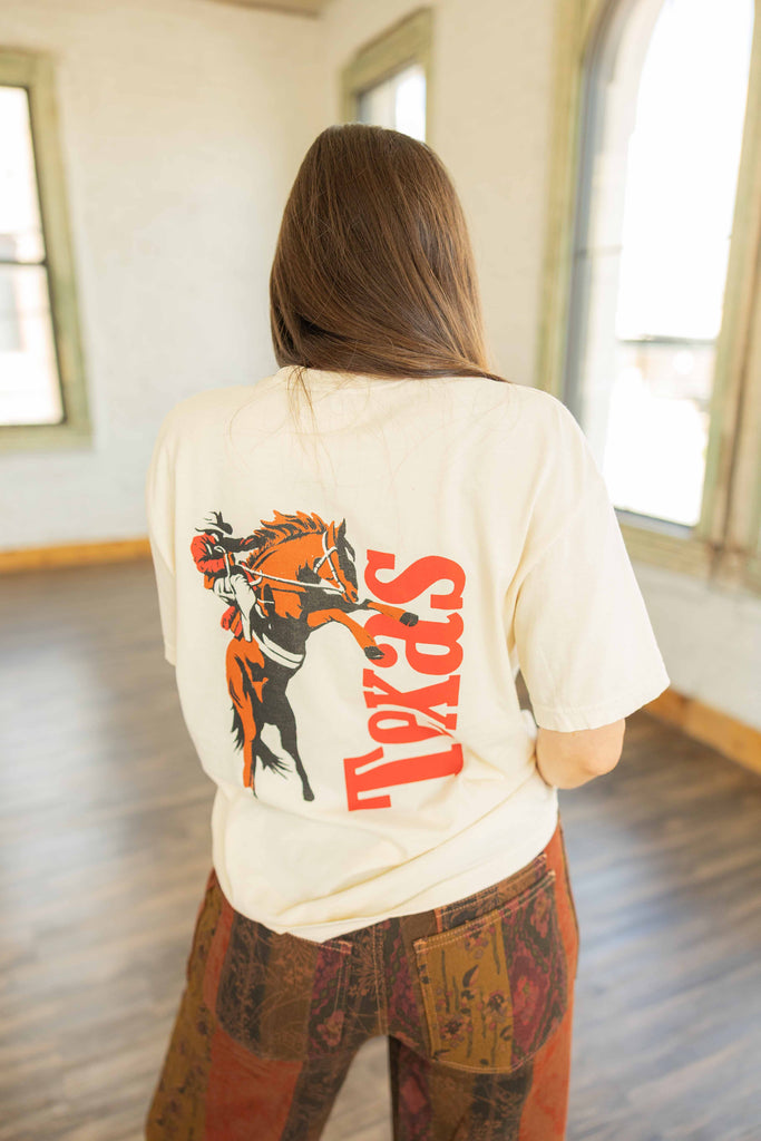 Kayla wearing the Texas Cowboy Icon graphic tee in cream with red Texas print on front and cowboy graphic on back, styled with printed pants inside Ruby and Yaya’s studio.