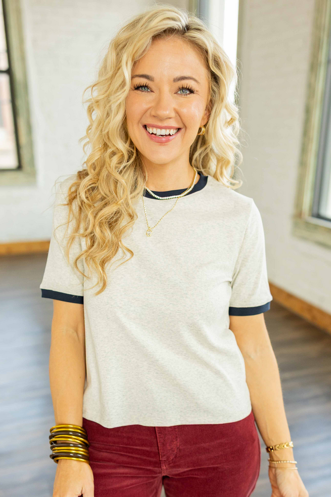 Holly wearing the Edith Ringer Everyday Tee, a light heather gray short sleeve top with navy trim on the collar and sleeves, styled with burgundy corduroy pants.