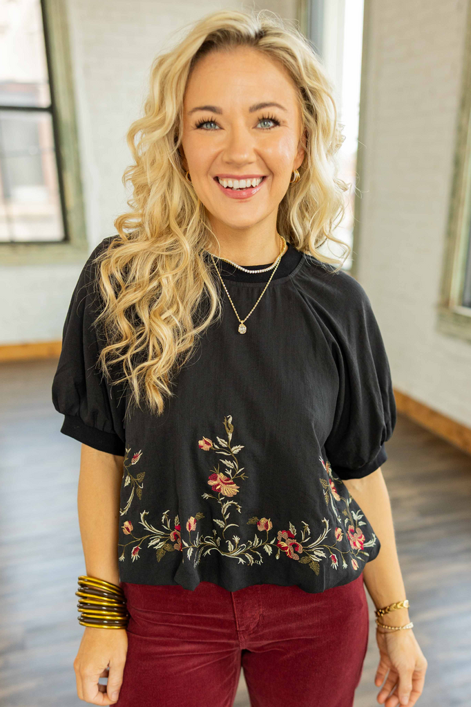 Holly wearing the Dayanna Embroidered Bubble Hem Waist Crop Top, a black short sleeve blouse with red and gold floral embroidery along the hem, paired with burgundy corduroy pants.