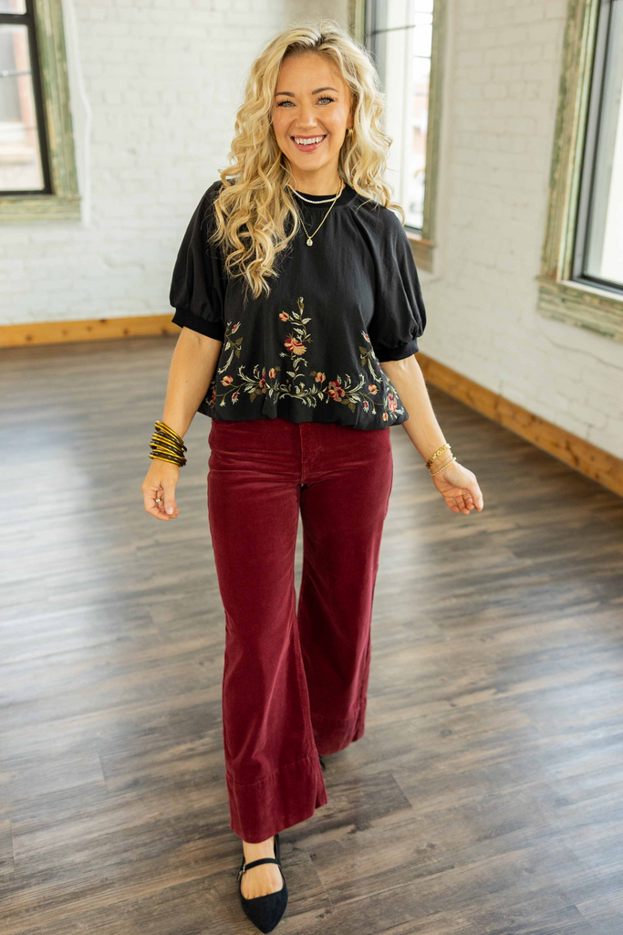 Holly wearing the Dayanna Embroidered Bubble Hem Waist Crop Top, a black short sleeve blouse with red and gold floral embroidery along the hem, paired with burgundy corduroy pants.