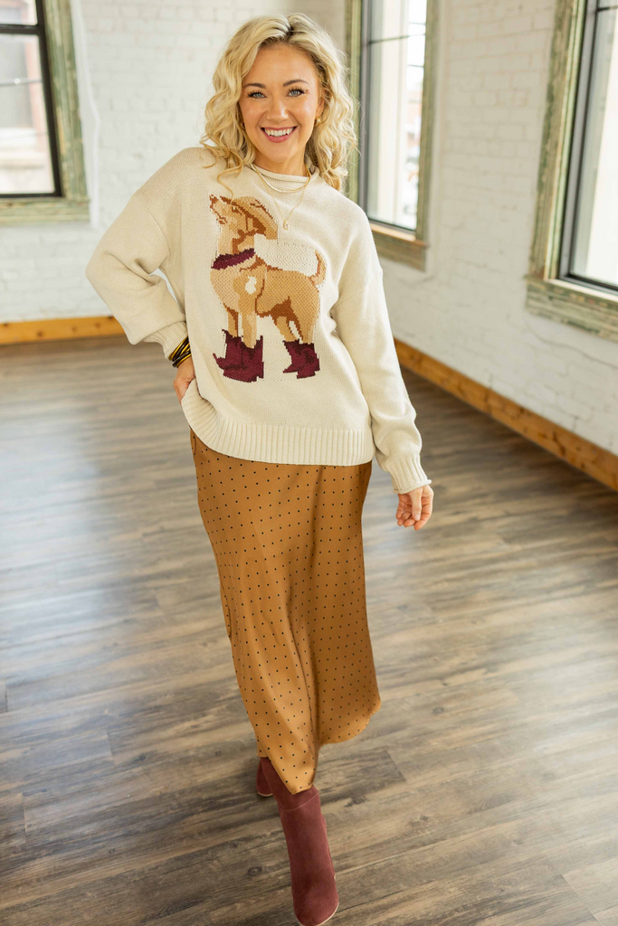 Holly wearing the Western Dog Sweater, a cream pullover sweater with a tan dog wearing a cowboy hat and maroon boots design, paired with a brown polka dot midi skirt.