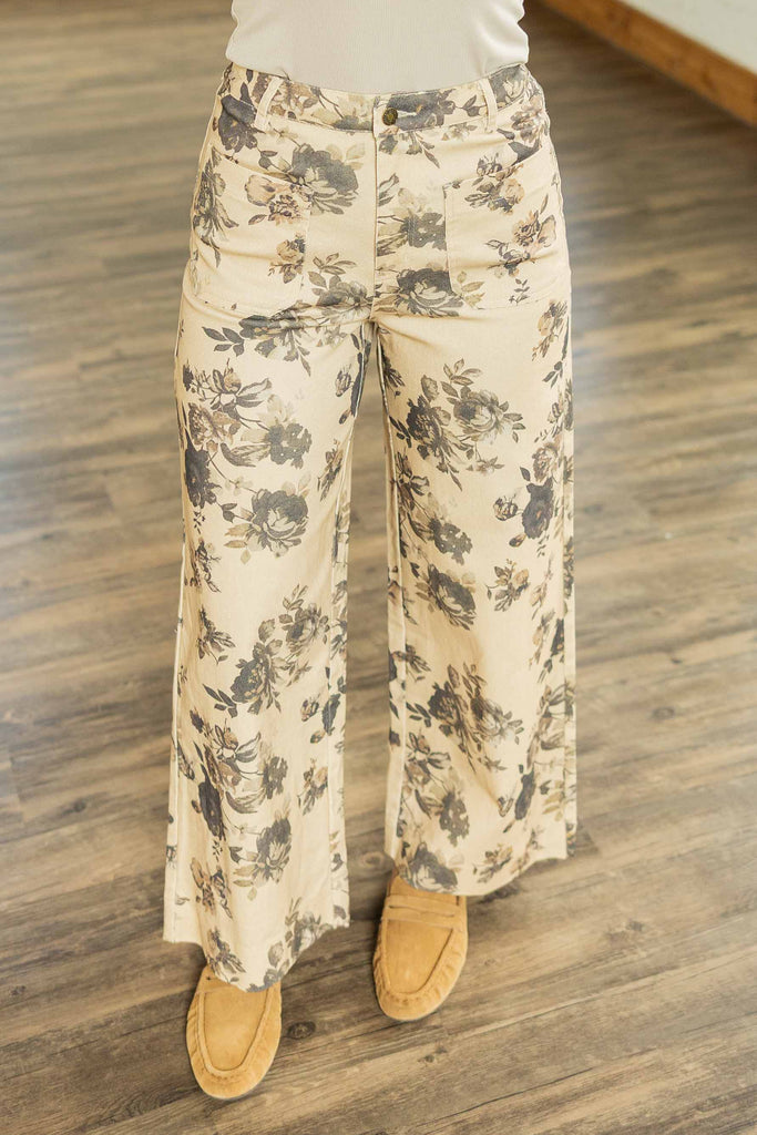 Holly wearing the Alicia Floral Cropped Pants, beige wide-leg cropped pants with charcoal floral print and front patch pockets, styled with a tan knit bow-tie vest top and loafers.