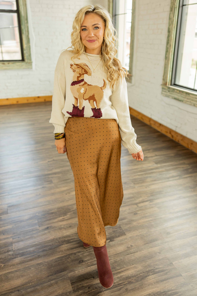 Holly wearing the Lana Satin Dot Midi Skirt, a camel-colored satin midi skirt with small black polka dots and an elastic waistband, styled with a cream sweater featuring a dog motif and burgundy boots.
