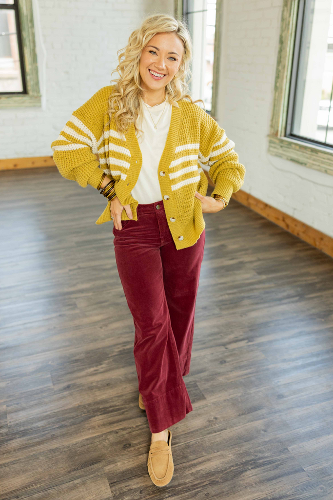 Holly wearing the Journey Striped Cardigan, a mustard yellow knit cardigan with off-white horizontal stripes and button front, styled with a white tee and burgundy corduroy pants.