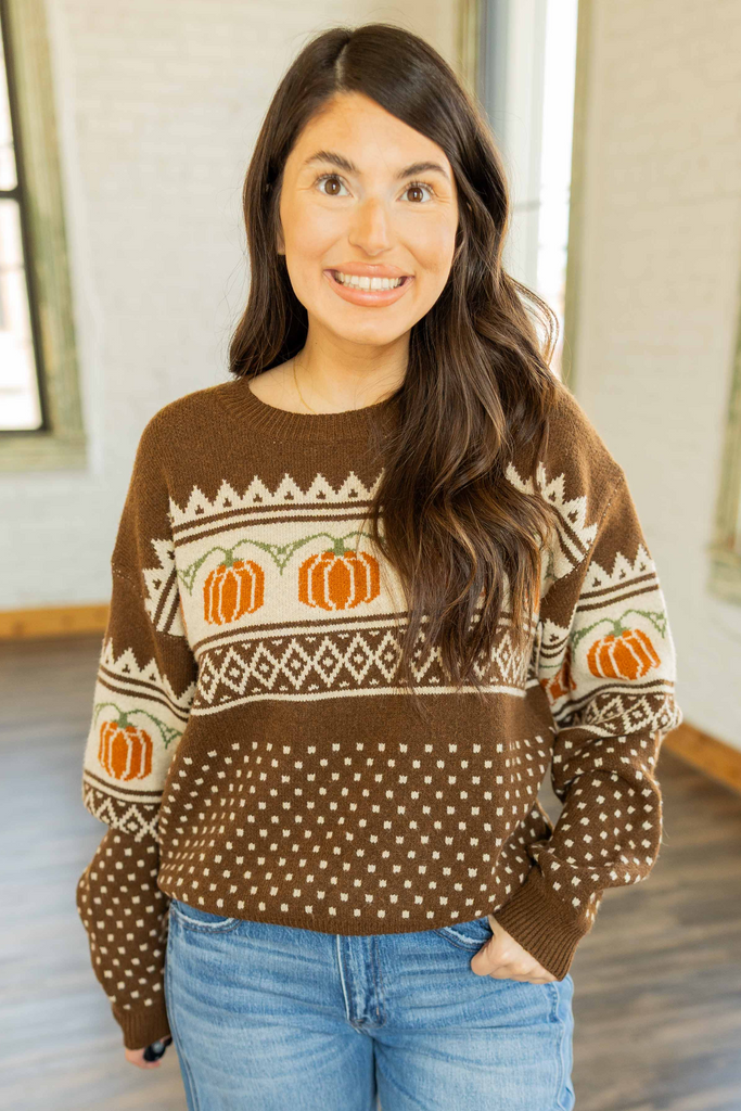 Kayla wearing the Oaklyn Pumpkin Sweater, a brown long sleeve knit sweater with cream and orange pumpkin jacquard pattern, styled with light wash wide leg jeans.
